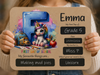 A person holds a personalized first-day-of-school sign featuring a cartoon unicorn, school supplies, and chalkboard-style text fields for the name Emma, grade 5, date, teacher, favorite thing, and future goal.