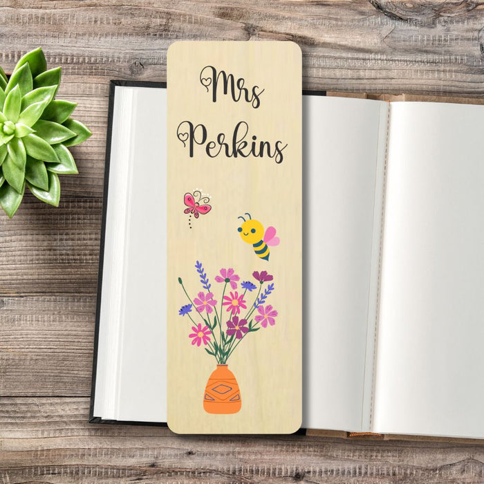 A personalized wooden bookmark with the text 'Mrs Perkins' and illustrations of a bee, butterfly, and flowers in a vase, displayed on an open notebook on a rustic wooden table next to a green succulent.