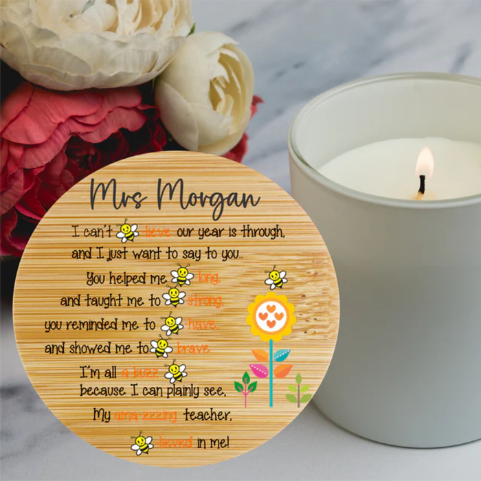 A personalized round wooden plaque with a printed message for a teacher, styled on a marble surface next to a lit white candle and flowers.