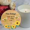 A personalized round wooden plaque or coaster with a printed thank-you message, two bumblebees, and yellow sunflowers, styled on a marble surface next to a lit candle and flowers.